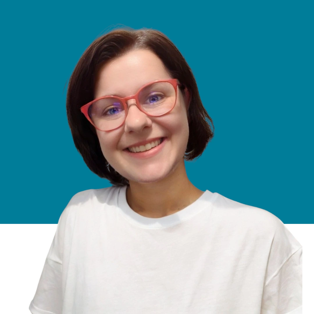 Marjorie, souriante, aux cheveux courts et châtain foncé, portant un t-shirt blanc uni. L’arrière-plan est divisé en deux : la partie supérieure est bleu-vert, la partie inférieure blanche.